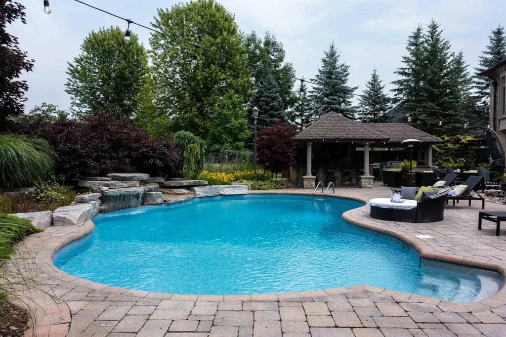 Curved in-ground pool with stone coping, a rock waterfall, and surrounding lounge area in a landscaped backyard.
